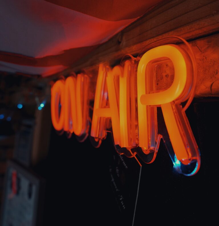 Illuminated orange 'On Air' neon sign hanging from a wooden ceiling in a radio studio