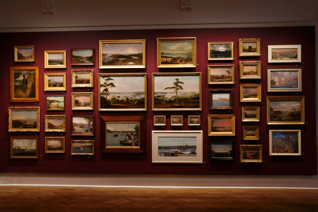 Gallery wall densely hung with framed landscape and portrait paintings in gold frames