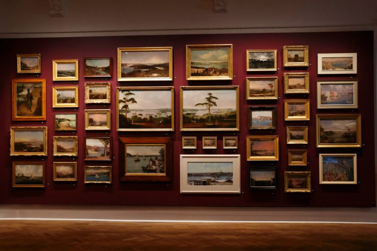 Gallery wall densely hung with framed landscape and portrait paintings in gold frames